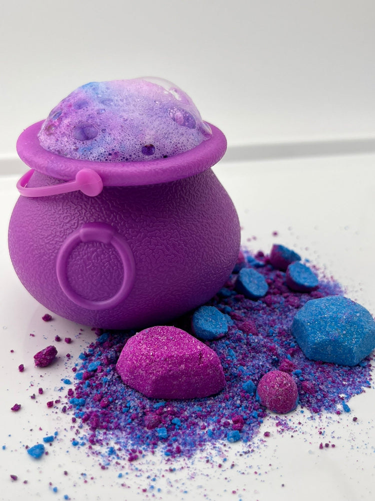 A photograph of Witch's Whirl potion making kit ingredients next to a purple cauldron, the cauldron is about to overflow with a foamy potion. 